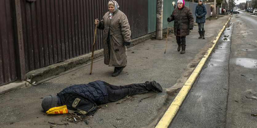 Leiche liegt auf dem Gehweg, alte Frauen gehen vorbei