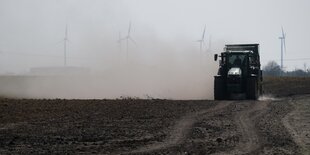 Traktor auf Acker in großer Staubwolke, im Hintergrund Windräder.