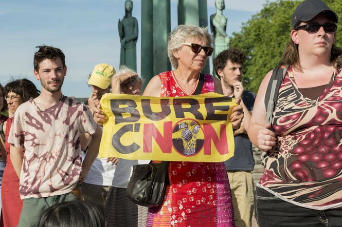 Eine ältere Frau mit einem Sommerkleid und Sonnebrille hält in einer Gruppe ein Protestplakat mit dem Namen des geplanten Endlagers Bure