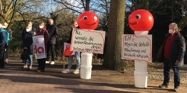 Bei einer Solikundgebung vorm Bremer Amtsgericht haben sich Leute versammelt. Zwei Demofiguren mit roten Köpfen tragen 2 Schilder: "Kennste die Senioren Residenz Gruppe Orpea? Soll ja Klagen geben", und: "Mögen sie deshalb Mitbestimmung nicht so sehr?"