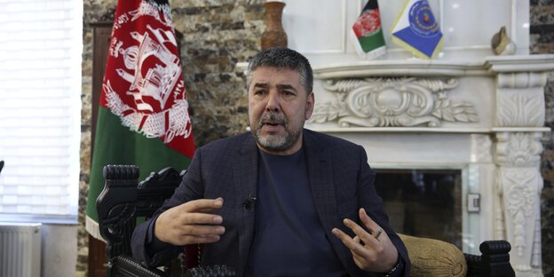 Ein Mann sitzt in einem Stuhl, im Hintergrund eine afghanische Flagge.