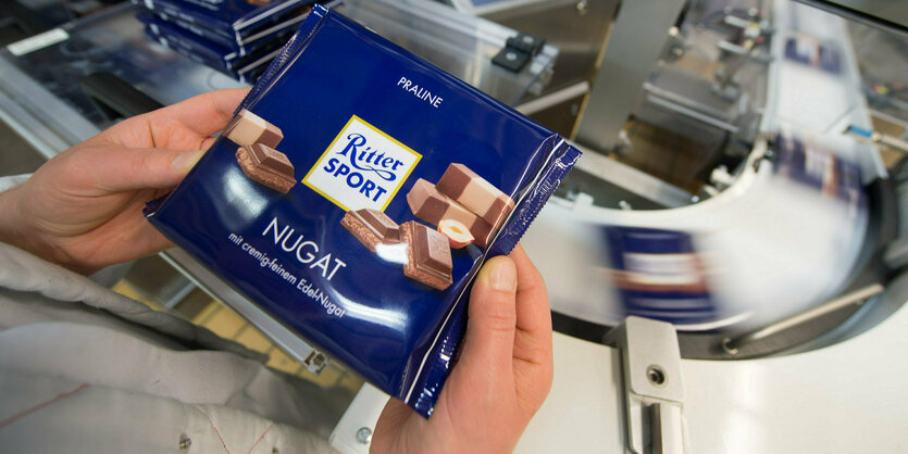 Zwei Hände halten eine Tafel Ritter Sport Nougatschokolade, im Hintergrund schießen andere Tafeln auf einem Laufband vorbei