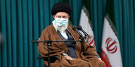 Ein Mann mit Maske und Turban sitzt auf einem Stuhl vor der iranischen Fahne.