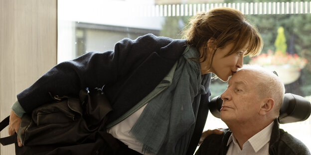 Emmanuèle gibt ihrem im Pflegestuhl sitzenden Vater André einen Kuss auf die Stirn.