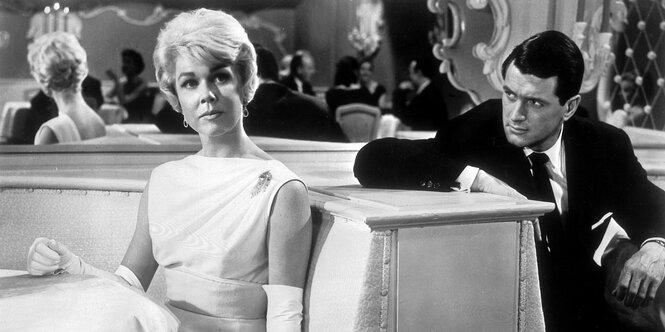 Doris Day sitzt in einem Restraurant, Rock Hudson sitzt daneben und schaut sie an.