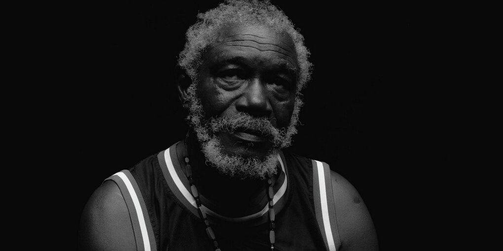 Schwarz-weiß Porträt von Horace Andy: Der Reggae-Musiker trägt ein ärmelloses Short und eine Kette und hebt den Blick nach oben