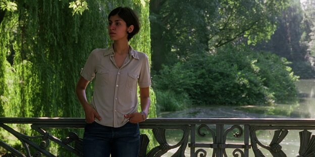 Eine junge Frau steht auf einer Brücke mit einem Park, die Sonne scheint im Hintergrund und ihre Hemdsärmel sind hochgekrempelt
