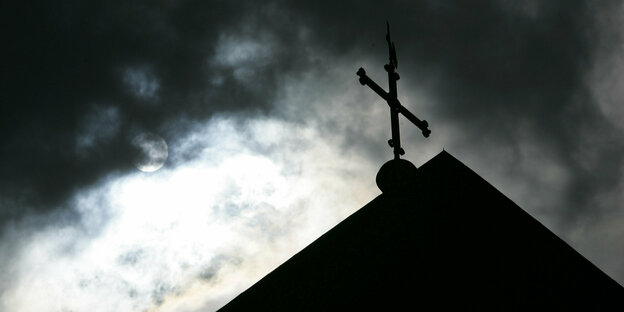 Im Gegenlicht und vor wolkenverhangenem Himmel ist die Kirchturmspitze eines Doms mit kreuz zu sehen