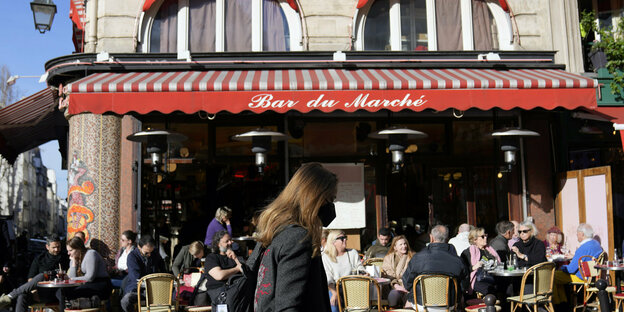 Eine Frau mit Maske spaziert an einem Cafe mit geöffnetem Außenbereich vorbei