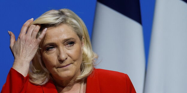 Eine blonde Frau mit rotem Oberteil blickt müde zur Seite, hinter ihr stehen französische Fahnen