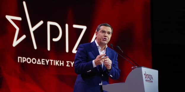 Vor einer beleuchteten Wand mit griechischen Buchstaben steht Alexis Tsipras und spricht in ein Mikrofon an einem Podest