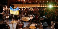 Die ukrainische Band Dakh Daughters von hinten auf einer Bühne, in den Händen die ukrainische Flagge