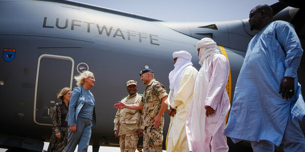 Eine blonde Frau und eine dunkelblonde Frau begrüßen zwei Soldaten und drei Männer in langen Roben vor einem Flugzeug auf dem "Luftwaffe" steht