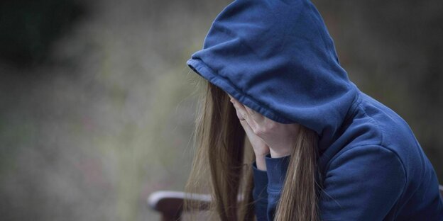 Eine junge Frau hat ihre Kapuze ins Gesicht gezogen, das sie mit ihren Händen bedeckt