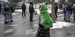 Eine Frau steht mit einer Blume in der Hand, einem grünen Tuch über dem Kopf im Schnee, umringt von Sicherheitskräfte, die sie beobachten