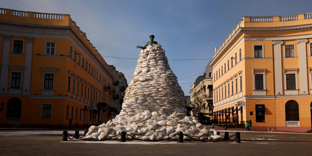 Ein Denkmal ist mit Sandsäcken geschützt, nur eine Kopf schaut heraus, die Häuser in der Altstadt leuchten gelb