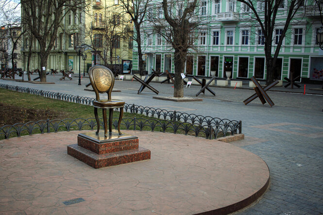 Ein riesiger Stuhl auf einem Denkmal, dahinter Panzersperren in einer Strasse