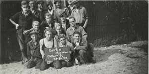 Eine Gruppe von Männern kniet im Sand mit einem Schild: Berlin Spandau Ostlager 5/1, 31-8-43
