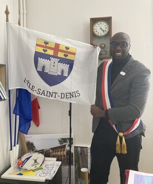 Ein Mann hat scih die Bürgermeisterschleife umgelegt und hält die Fahne von I `Ile Saint-Denis in der Hand