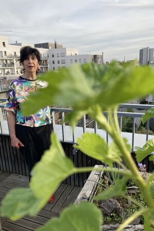 Eine Frau steht hinter einer hohen grünen Pflanze auf einem Balkon, dahinter Hochhäuser