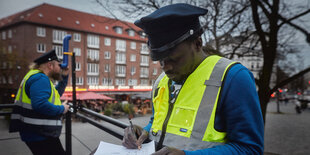 Ein Mann in gelber Weste und mit einer Polizeimütze blickt durch ein Fernrohr über den Hansaplatz in Hamburg. Daneben steht ein anderer Mann in gelber Weste und mit Polizeimütze, der etwas in einen Notizblock schreibt