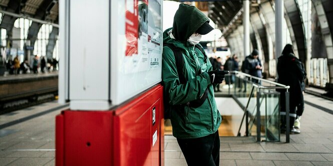 Ein Mann trägt eine grüne Jacke und eine Mundschutzmaske, er schaut auf sein Handy