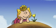 Sally, Charlie Browns Schwester aus der Comicreihe "Die Peanuts" liegt auf dem Bauch auf einem Erdhügel und schaut lächelnd einer gelben Blume beim Wachsen zu