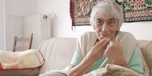 Eine alte Frau sitzt auf einem Sofa, sie benutzt die Hände während sie lächelnd erzählt