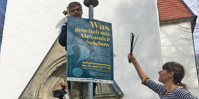 Zwei Personen hängen ein Plakat auf. Darauf wird die Auftaktveranstaltung des Gedenkprojekts angekündigt.