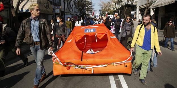 Menschen tragen ein rangefarbenes Rettungsfloß durch die Straßen von Athen