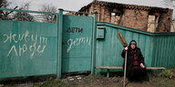 Eine ältere Frau sitzt vor einem Zaun, mit der Aufschrift "Hier leben Menschen" auf Russisch, dahinter ein zerbombtes Haus