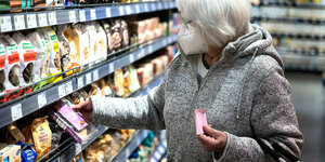 Eine ätere Dame holt in einem Supermarkt etwas aus dem Regal, dabei trägt sie eine Maske