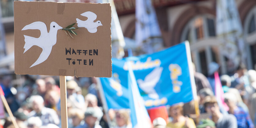 Schild mit Friedenstaube und Aufschrift "Waffen töten" vor unscharf fotografierter Demonstration