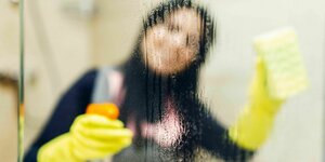 Eine Frau mit gelben Handschuhen putzt eine Duschkabine, man sieht sie verschwommen durch das Glas