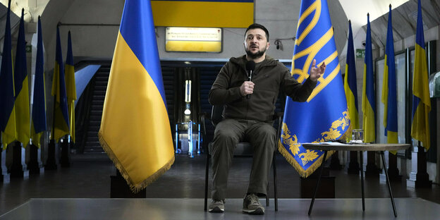 Präsident Selenski sitzt zwischen ukrainischen Fahnen und spricht ins Mikrofon