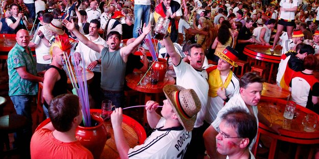 Das Innere vom Bierkönig. Ungezählte Menschen stehen neben und auf Tischen. Sie trinken Alkohol, tanzen und grölen. Viele von ihnen tragen Deutschlandtrikots oder anderen Merchandise der deutschen Nationalmannschaft wie etwa riesige Hüte.
