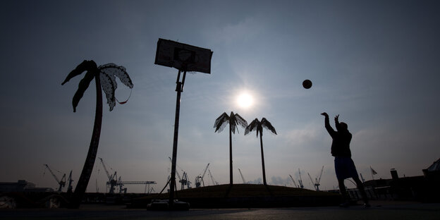 Ein Mann wirft in der untergehenden Sonne in einem leeren Park mit künsltichen Palmen einen Ball auf einen Basketballkorb