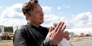Elon Musk, ein mittelaltermann mit kurzen, fluffigen Haaren. Er grinst in die Kamera und hebt seine Hände wie zum Gebet. Im Hintergrund die Brache von Grünheide.