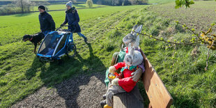 Ein Paar mit Hund und Kinderwagen geht an 2 Osterhasenfiguren, die auf einer Bank sitzen vorbei