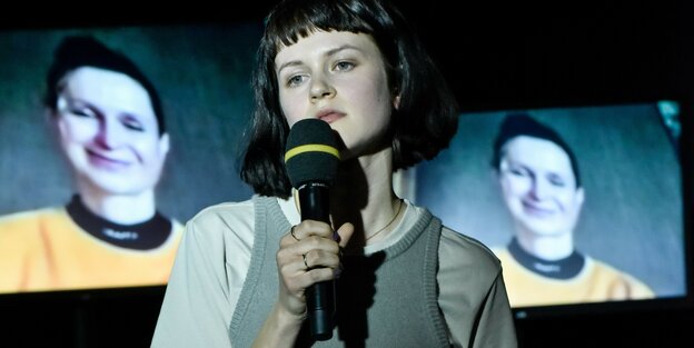 Die junge Schauspielerin Sidney Fahlisch hält auf der Bühne ein Mikro in der Hand, hinter ihr, als Video, Manuela Fahlisch.