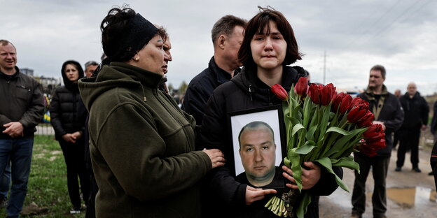 Ein trauerndes Mädchen hält ein Foto ihres toten Vaters und einen roten Tulpenstrauß in der Hand, sie wird von einer Frau getröstet
