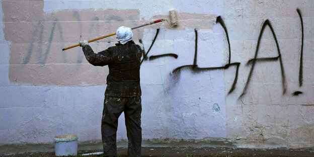 Eine Pesron in Malerklamotten und Farboroller übermalt mit weißer Farbe Graffitti-Schrift an einer Wand