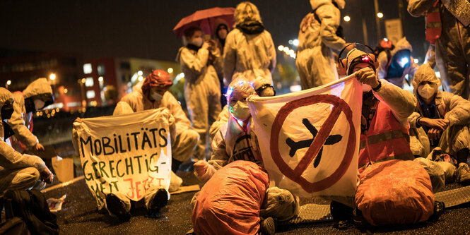 Protestierende in weißen Schutzanzügen, ein Schild mit einem durchgestrichene Flugzeug