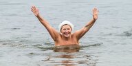 Eine Frau mit Bademütze steht bis zu den Brüsten im Wasser und winkt mit beiden Armen. Ihr Oberkörper ist nackt