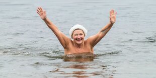 Eine Frau mit Bademütze steht bis zu den Brüsten im Wasser und winkt mit beiden Armen. Ihr Oberkörper ist nackt