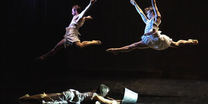 Zwei Tänzer springen hoch durch die Luft, eien Tänzerin liegt unter ihnen auf der Bühne am Boden und schaut in einen Eimer aus Metall