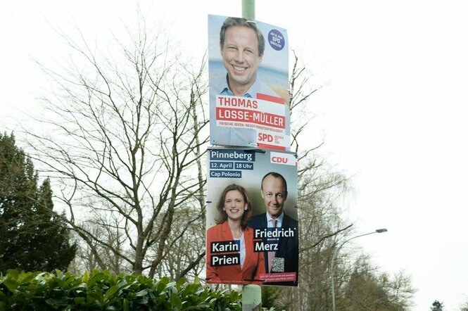 Wahlplakate mit den Portraits von Karin Prien und Friedrich Merz hängen unter einem SPD Plakat an einer Laterne