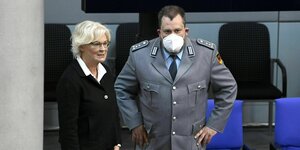 Christine Lambrecht seht im Bundestag neben einem Mann mit UNiform. Sie wirkt gestresst, er stemmt forsch die Hände in die Hüften, aber man sieht seinen Gesichtsausdruck nicht, wegen Mund-Nasen-Schutz
