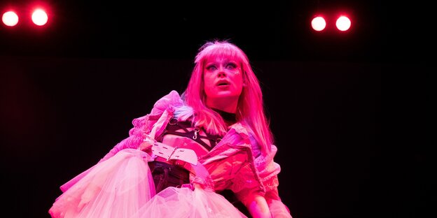 Eine Schauspielerin in einem Kleid steht etwas schief auf einer dunklen Bühne und wird rosafarben angestrahlt