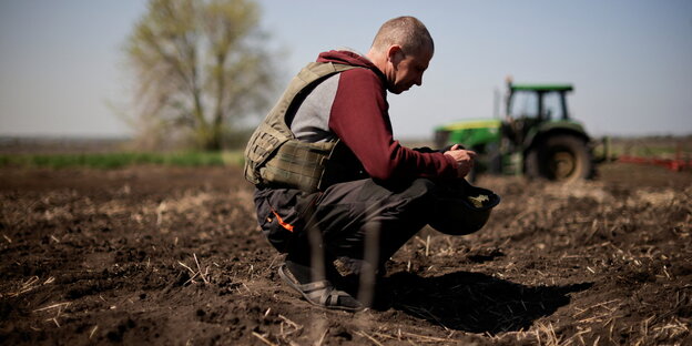 Ein Landwirt mit schussischerer Weste kniet auf seinem Feld und bearbeitet den Boden